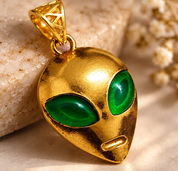 Close-up of the Astra Enigma alien pendant showing vivid green eyes, textured gold-tone finish and openwork bail