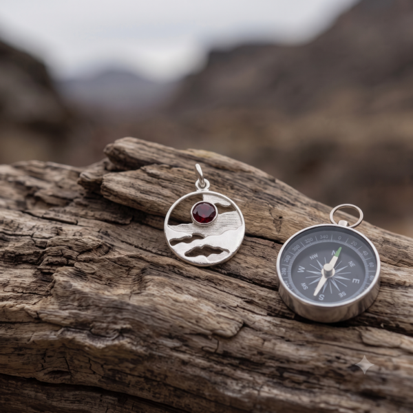 Horizon Solstice sterling silver landscape medallion with Ruby Red CZ beside a compass on weathered wood